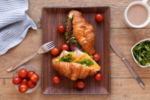 Flaky croissant egg sandwich with scrambled eggs, melted cheese, and crispy bacon, served on a wooden plate with a side of fresh fruit.