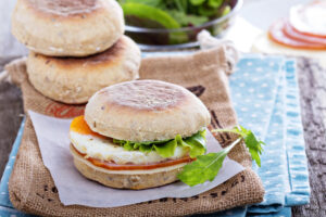 A perfectly toasted English muffin sandwich with a fried egg, melted cheese, crispy bacon, and fresh spinach on a wooden plate.