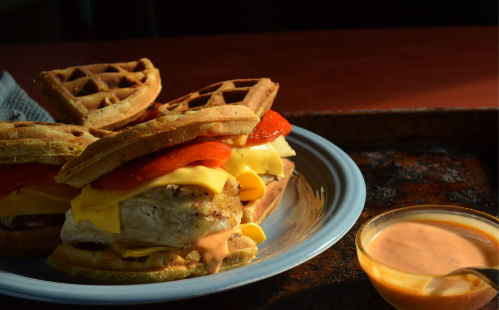 Chicken and waffle sandwich with spicy honey and fresh greens on a wooden plate.