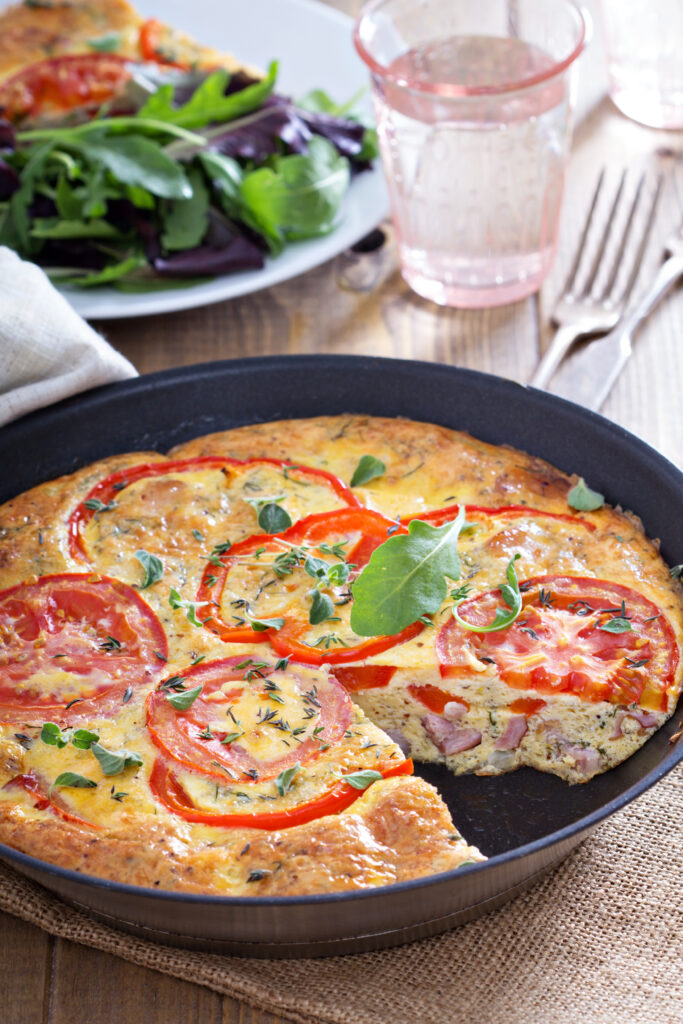 A fluffy ham and cheese frittata in a cast-iron skillet, topped with melted cheese and garnished with fresh parsley, served with toast and a side salad.