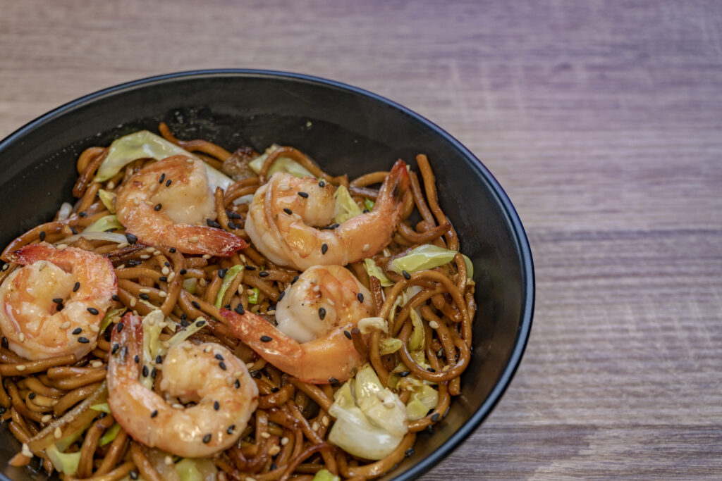 A bowl of shrimp chow mein with stir-fried vegetables, juicy shrimp, and golden noodles, garnished with green onions and sesame seeds.