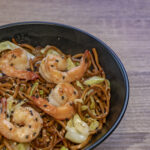 A bowl of shrimp chow mein with stir-fried vegetables, juicy shrimp, and golden noodles, garnished with green onions and sesame seeds.